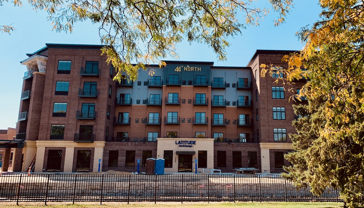 A five-story building with brick facade, with four floors of balonies. Near the center-top of the building a label reads 44° North. Below on the ground level is a sign reading Latitude Bar & Lounge. The building is nearly complete, at least the part of the exterior you can see,although a small bit of white housewrap is visible. The parking lot is still unpaved, and some construction materials and two portable toilets are in front of the building.